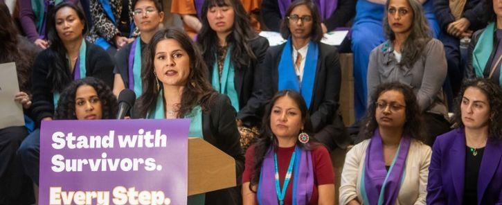 Councilmember Rinck speaking at a podium with a sign that reads "Stand with Survivors. Every Step.", many people in the background wearing orange, purple, and teal