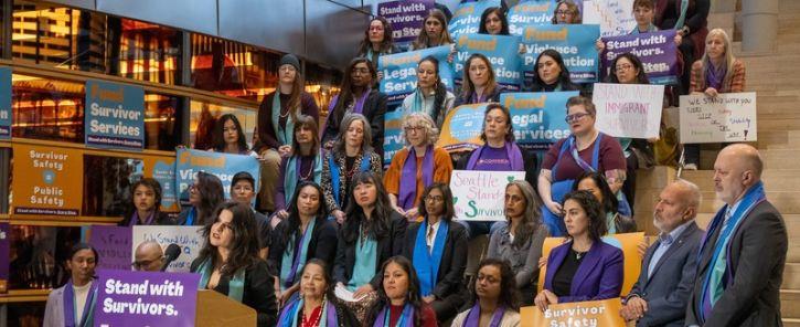 Councilmember Rinck speaking at a podium, many people sit on the stairs behind her and are wearing orange, purple, and teal