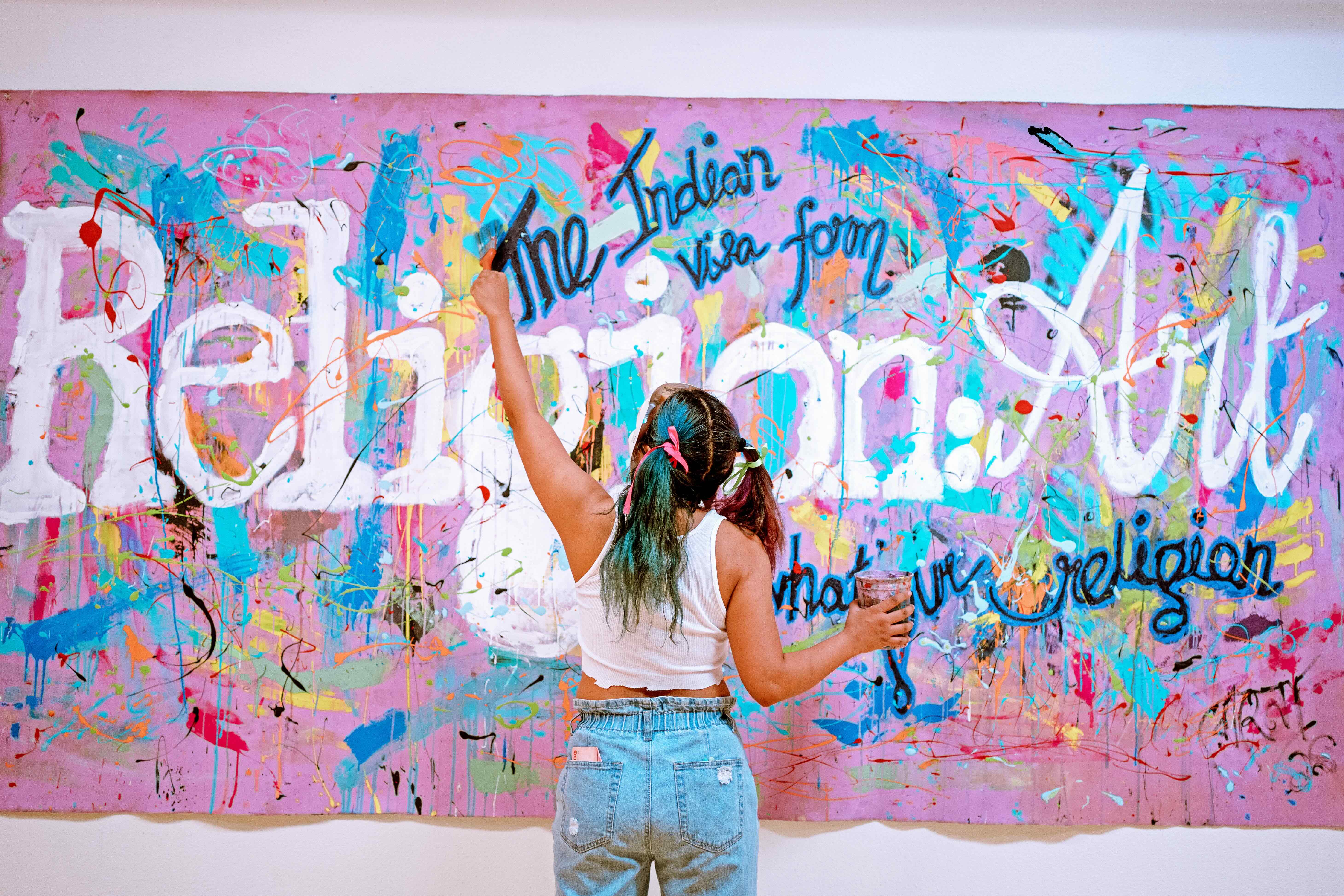 An artist works on a large canvas with her back to us. Her left arm is fully extended while holding a brush.