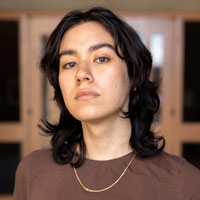 A young adult with dark shoulder length hair, wearing a brown tee shirt and gold necklace with a serious expression