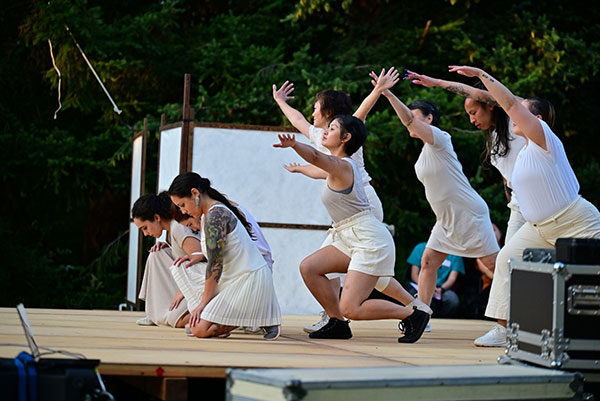 Eight dancers in white crouch on stage. Three in front down on their knees, five behind with their arms outstretched.