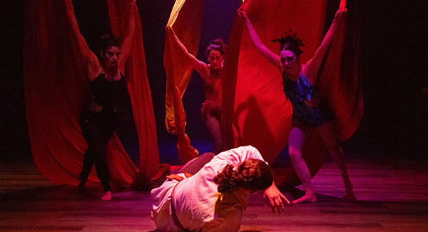 A man cowers onstage as three women lean toward him, arms extended upward to grip fabric.