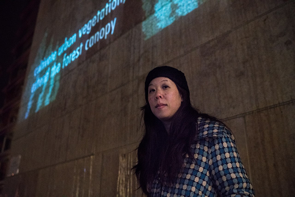 Civic Poet Shin Yu Pai, an Asian American with long black hair wearing a beanie, poses with an outdoor poetry projection
