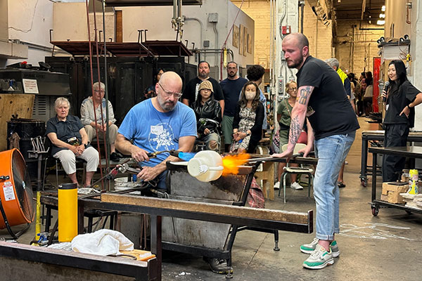Dave holds a torch to a piece of glass that's held by an assistant. A group of people look on.