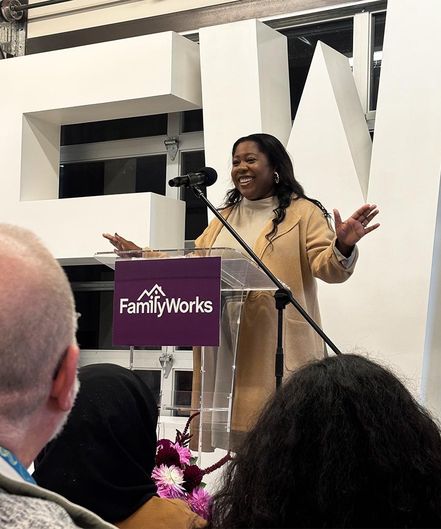 A smiling woman stands at a podium with her hands extended in welcome.