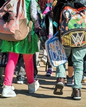 A group of young students wearing brightly colored backpacks walks down the sidewalk