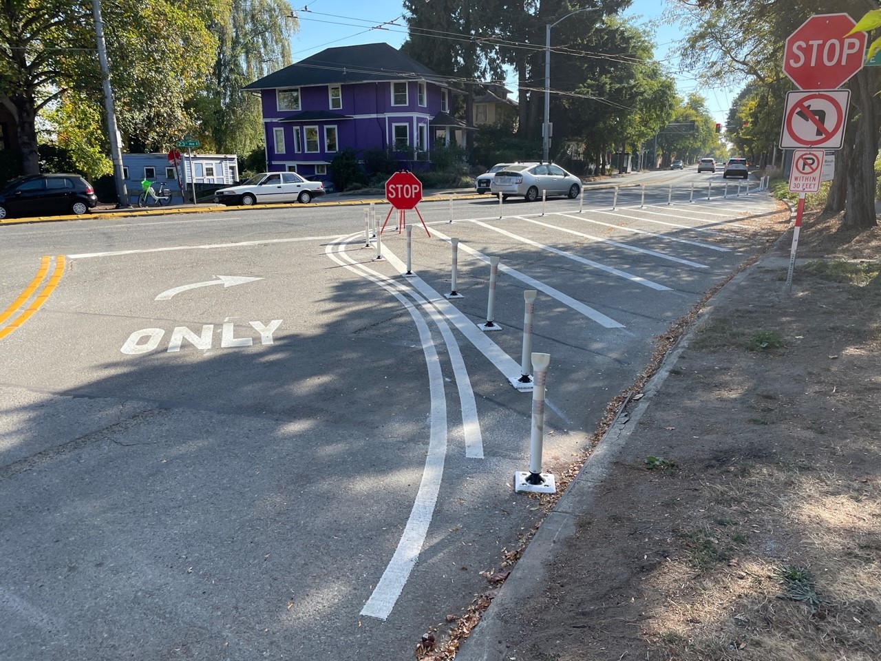 A picture showing a street with curved white lines painted and white posts in the street and a stop sign.