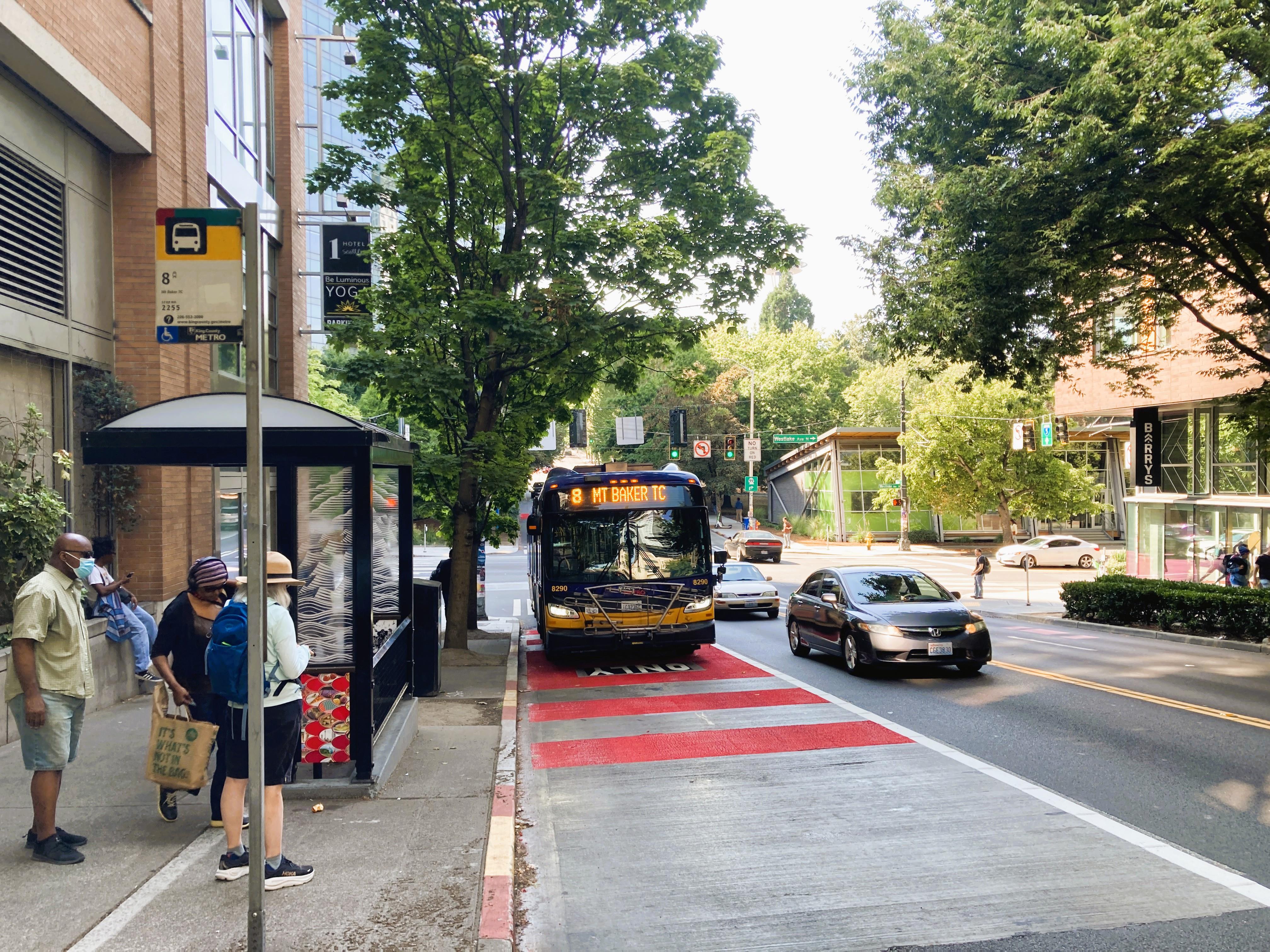 A King County Metro bus on Route 8 pulls into a vibrant red transit lane next to a modern bus shelter on Denny Way. 