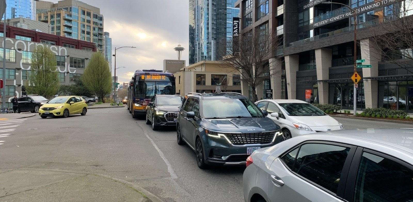 Traffic on Denny Way in Seattle, with several cars queued in the right lane and a Route 8 bus (Mt. Baker TC) behind them. Mid-rise buildings line the street, and the Space Needle is visible in the background under a cloudy sky.