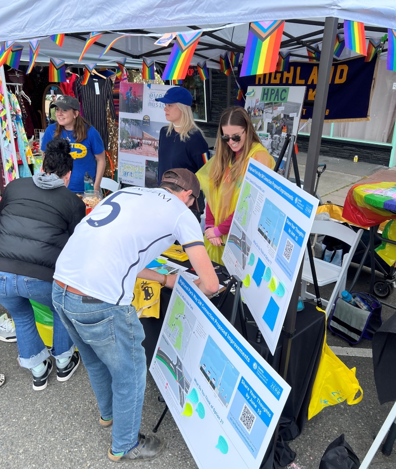 Woman standing behind a table with project materials on it. She is smiling and talking to a person who is writing a comment about the project. They are standing under a tent decorated with rainbow flags. There are poster boards on easels illustrating the different project design options.