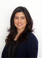 Dhyana smiling broadly at the camera in a dark blazer in front of a light background.