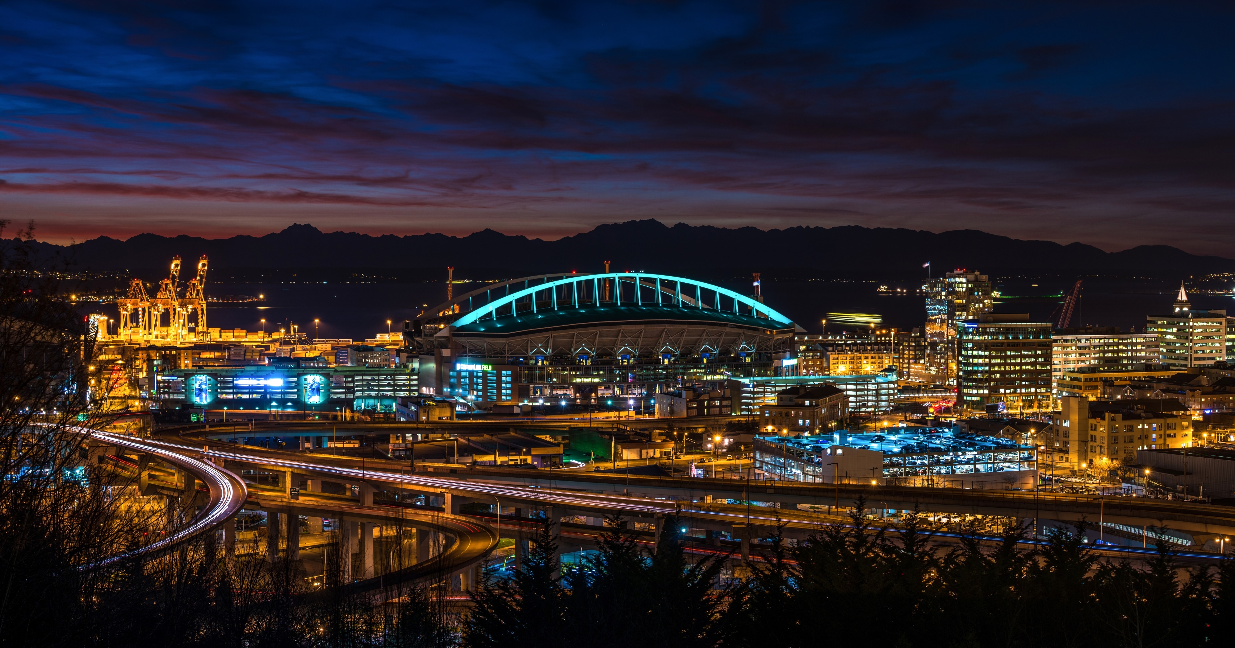 Nighttime view of a brightly lit stadium, surrounding highways with light trails, and nearby city buildings against a dark sky with colorful sunset clouds.
