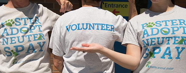 Three people wearing Seattle Animal Shelter shirts; two shirts read ‘Foster Adopt Neuter Spay’ and one reads ‘Volunteer.’