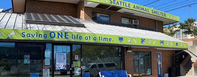 Entrance of the Seattle Animal Shelter with a green banner reading ‘Saving ONE life at a time’ and a blue bench in front.