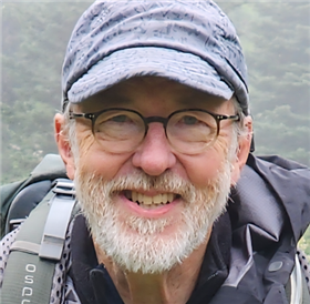A headshot of Review Panel member Bruce Flory. An outdoor portrait of Bruce wearing a light gray cap and a dark jacket. The background shows a misty, forested landscape with soft green trees.