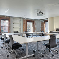 Large tables and rolling chairs are arranged in a square. A food area with a sink is on the side and wood-framed windows let in lots of natural light.