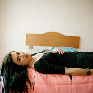 A young person lays on a bed, long hair draped off the edge, staring into the camera.
