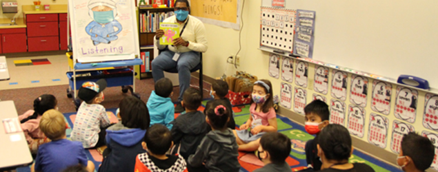 a pre-k classroom, kids sitting on the floor listening to a teacher read them a book