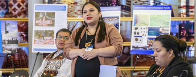 Image of an indigenous woman standing at a microphone speaking to a group