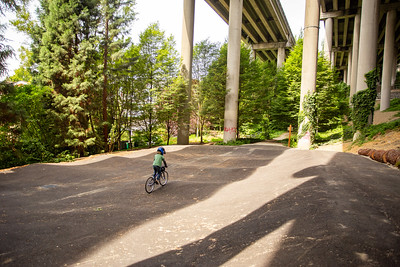 A child pedals a bike along paved pump track under the I-5 Highway, surrounded by trees and freeway supports overhead