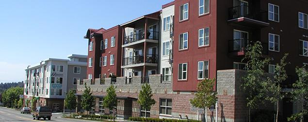 A row of modern multi‑story apartment buildings along a street, with trees and parked cars in front.