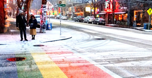 People about to cross a street with snow.  The crosswalk is a rainbow crosswalk. 