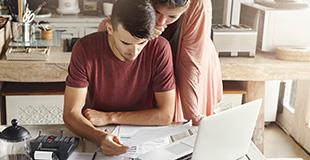 a woman leans over a man who is sitting at a desk with paperwork and a laptop