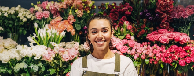 Florist in apron smiling and standing in front of flowers with arms crossed.
