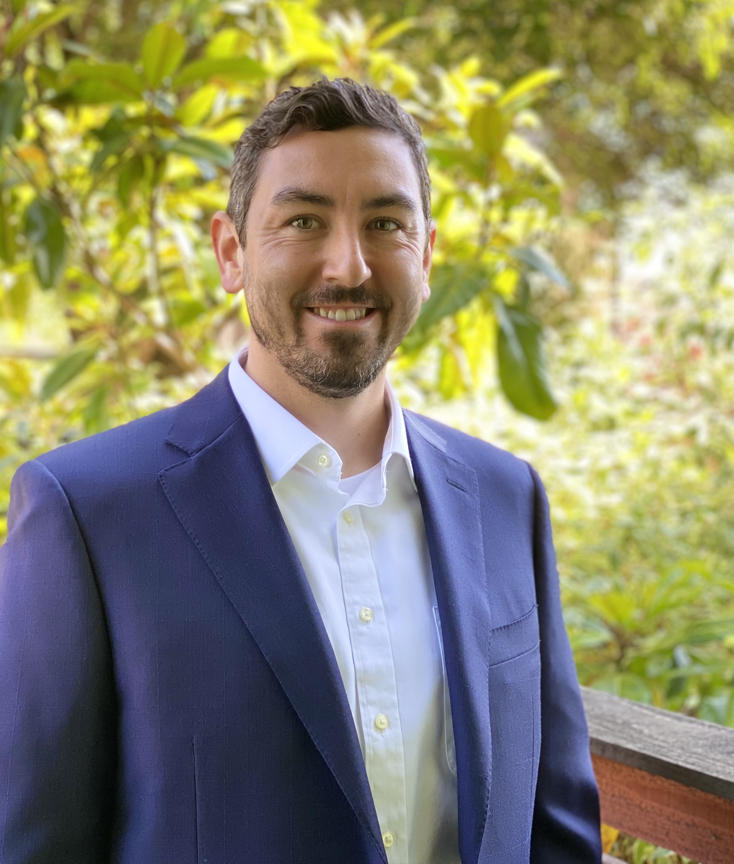 Portrait of Nicholas smiling broadly at the camera in a blue blazer with green foliage in the background.